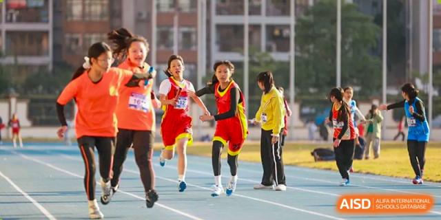 Students from Humen Foreign Language School International Division in athletic gear run a relay race on a blue track outdoors during a sports event.