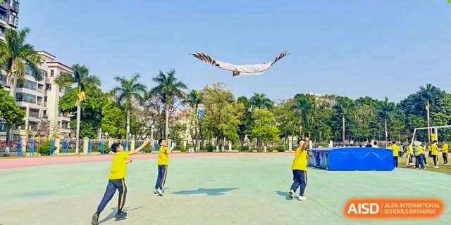 Students from Humen Foreign Language School International Division play on a green sports field while a large bird, possibly an eagle, flies overhead in the sunny sky.