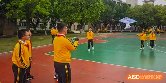 Students from Humen Foreign Language School International Division in yellow shirts are on an outdoor basketball court, with one student holding a ball, practicing sports.
