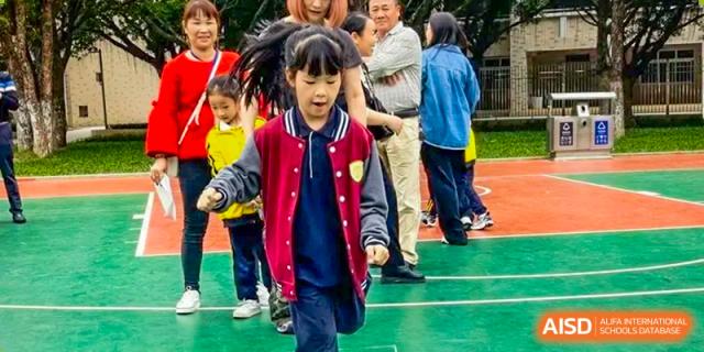 A young female student from Humen Foreign Language School International Division runs happily on a grassy area, with other students and adults watching in the background.