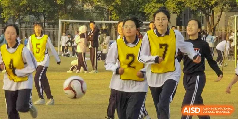 Female students from Humen Foreign Language School International Division wearing sports jerseys participate in a game of soccer or football on an outdoor field.