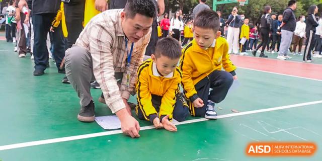 A teacher from Humen Foreign Language School International Division assists two young students in yellow shirts with an outdoor activity on a green and red court.