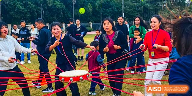 Adults and students from Humen Foreign Language School International Division participate in a team-building activity involving a large drum and multiple ropes.