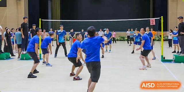 Players from Humen Foreign Language School International Division compete in a game of indoor volleyball, with one player in a blue shirt ready to receive the ball.
