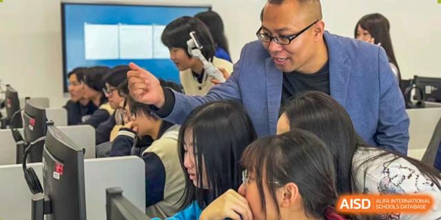 A teacher at Maple Glory Education leaning in to help a female student with an assignment in a busy classroom setting.