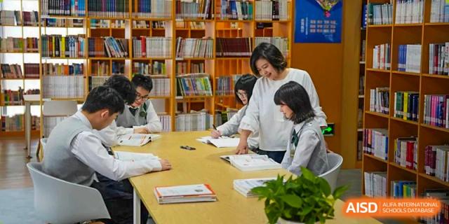 A female teacher at Maple Glory Education assisting students with their work at a table surrounded by bookshelves in a library or study area.
