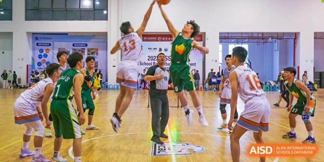 An action shot of two Maple Glory Education players jumping high for a rebound during a basketball game, with a referee watching.