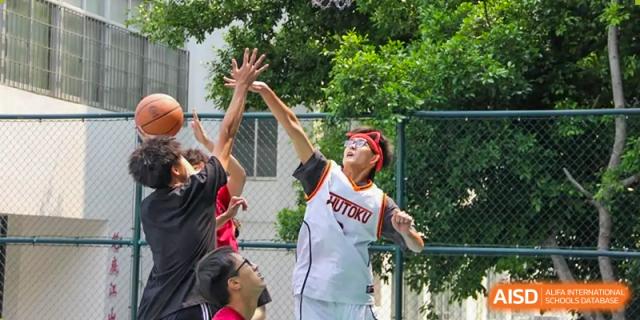 Two Maple Glory Education players, one in a white and red jersey, reaching up to block a shot on an outdoor basketball court.