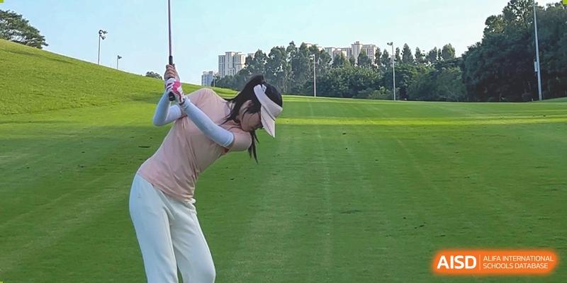 A Maple Glory Education student playing golf, swinging a club on a green course with trees in the background.
