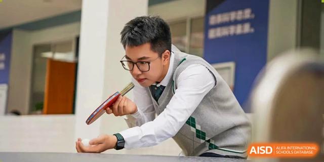 A male student from Maple Glory Education wearing glasses, a shirt, and a gray vest, intensely focused on a table tennis match.