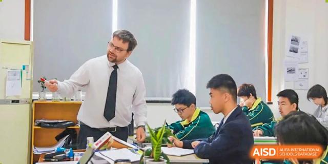 A male teacher in a white shirt and tie helping two male students with their work at a desk in a classroom at Maple Leaf International School Wuhan.