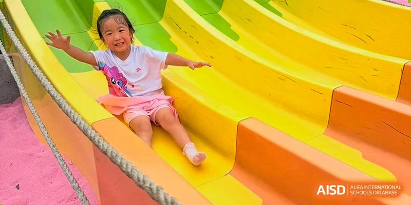 A young girl smiling brightly while sliding down a bright yellow and orange slide on the playground at Maple Leaf International School Wuhan.