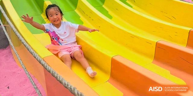 A young girl smiling brightly while sliding down a bright yellow and orange slide on the playground at Maple Leaf International School Wuhan.