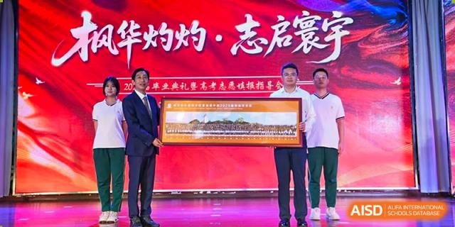 Two male school officials holding a large framed photo of a graduating class on a stage with a red backdrop at Maple Leaf International School Wuhan.