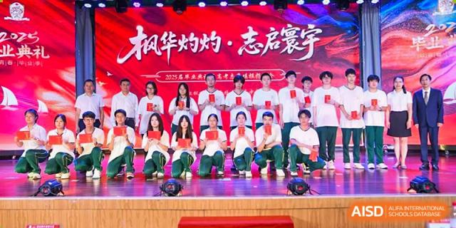A group of male and female students in white and green shirts kneeling and standing on a stage, holding red envelopes or awards, at Maple Leaf International School Wuhan.