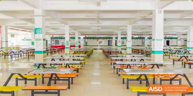 A view of the large, brightly lit and empty school cafeteria or dining hall, featuring rows of tables and benches with colorful orange and yellow seating at Maple Leaf International School Wuhan.