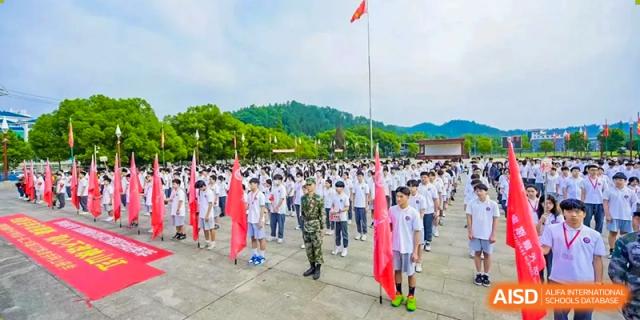 A large student assembly outdoors for a flag-raising ceremony or training, with many students holding red flags and a Chinese flag flying overhead at Oxstand International School, Shenzhen.