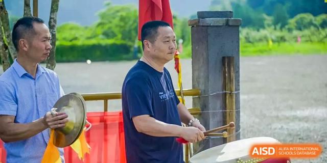 A male instructor in a blue shirt playing a drum outdoors during an assembly or cultural event at Oxstand International School, Shenzhen.