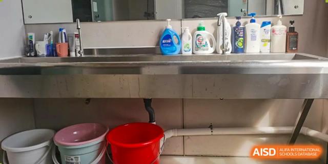 A close-up view of a metal utility shelf with cleaning supplies and a red bucket underneath in a service area at Oxstand International School, Shenzhen.