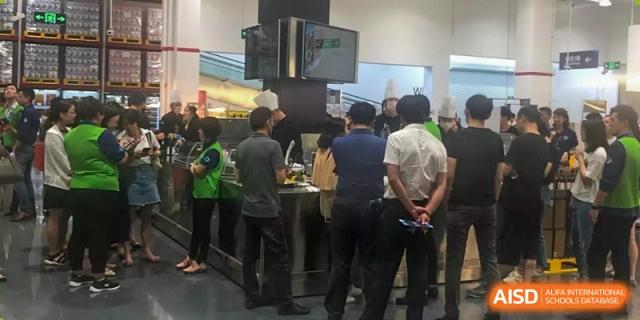 Adults gathered in a cafeteria or food service area at Shanghai Livingston American School, possibly for an event or food demonstration.