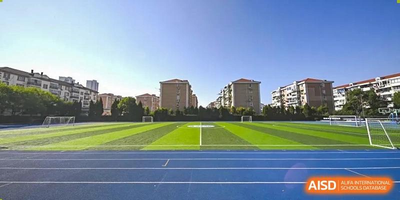 A wide shot of the outdoor sports field with a green artificial turf area and a blue running track on a sunny day at Shanghai United International School (SUIS-Jiaoke) Jiaoke Campus.