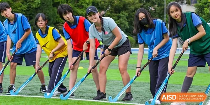 A coach or student leader from Shanghai United International School (SUIS-Jiaoke) Jiaoke Campus demonstrating a technique with a field hockey stick on an outdoor turf field, surrounded by students.