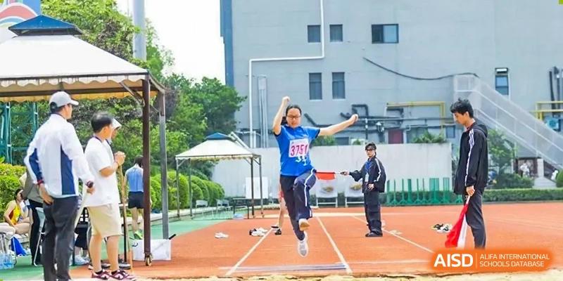 A student in a blue and red sports uniform performing a long jump during a sports day event at Shanghai United International School (SUIS-Jiaoke) Jiaoke Campus.