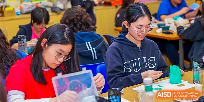 Close-up of students working on tablets or laptops, one wearing a 'SUIS' hoodie, in a classroom or study area at Shanghai United International School (SUIS-Jiaoke) Jiaoke Campus.