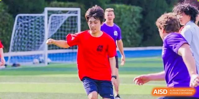An athlete in a red shirt and dark shorts playing a field sport, possibly soccer or rugby, at Shanghai United International School (SUIS-Jiaoke) Jiaoke Campus.