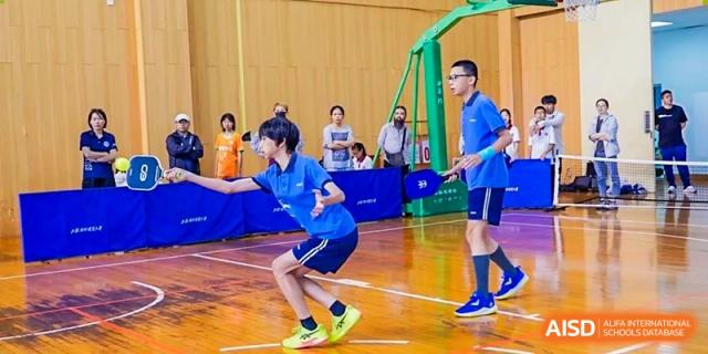 A student in a blue sports uniform playing table tennis with a high-energy stance in the gymnasium at Shanghai United International School (SUIS-Jiaoke) Jiaoke Campus.