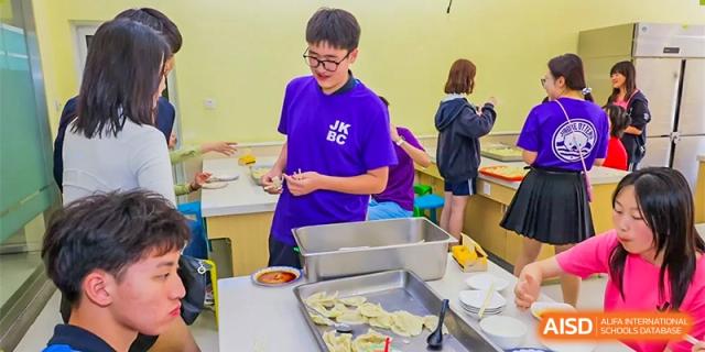 A male student in a purple shirt preparing food, possibly dumplings, on a tray in a kitchen or cafeteria line at Shanghai United International School (SUIS-Jiaoke) Jiaoke Campus.