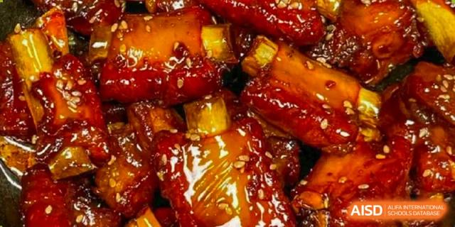 A close-up of a dish of glazed or braised pork ribs sprinkled with sesame seeds, representing a school meal or culinary offering at Shanghai United International School (SUIS-Jiaoke) Jiaoke Campus.