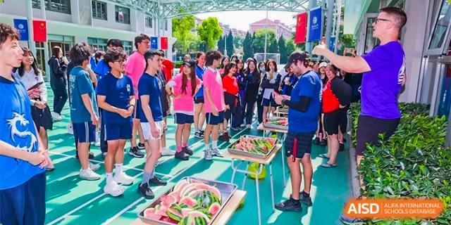 A group of students and teachers standing on a sports field, with tables of fresh watermelon slices for refreshment during an outdoor event at Shanghai United International School (SUIS-Jiaoke) Jiaoke Campus.