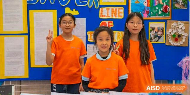 Two female students in bright orange shirts smiling behind a project display at a school fair or exhibition at Western International School of Shanghai.