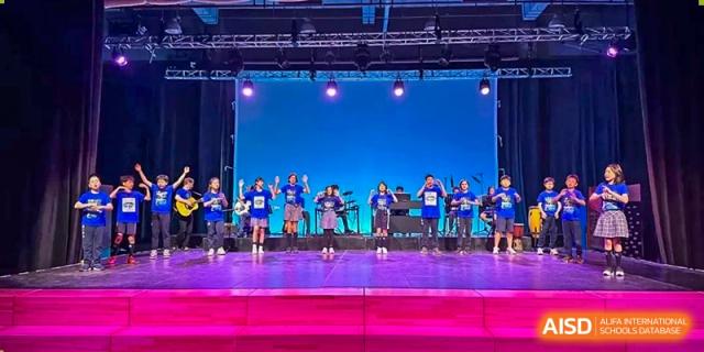 A group of students performing a band or dance routine on a brightly lit stage outdoors during a school festival at Western International School of Shanghai.