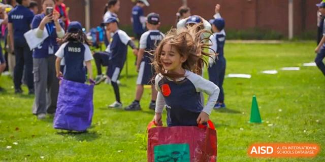 A happy young student sitting on a red block or cushion on a sports field during an activity at Western International School of Shanghai.