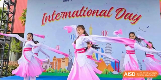 A female student performing a traditional dance in a pink and white costume with ribbons in front of a colorful "International" banner at Western International School of Shanghai.