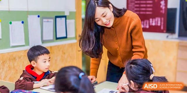 A smiling female teacher in an orange sweater leans over a desk to assist a young student with their work in a close-up shot at Yinghua Academy of Tianjin.