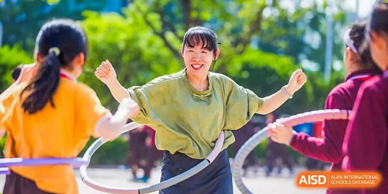 A smiling young woman in a green shirt doing an activity with a pink hula hoop outdoors at Yinghua Academy of Tianjin.