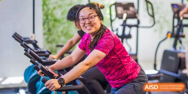 A smiling female student in a pink t-shirt and glasses exercises on a stationary bike in the school gym or fitness center at Yinghua Academy of Tianjin.