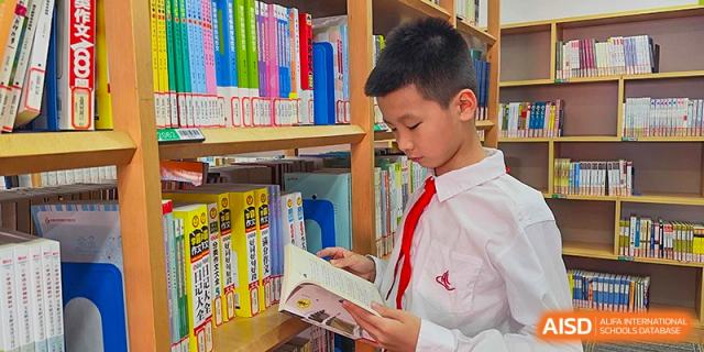 A male student in a white shirt and red tie stands next to a bookshelf, reading a book in the library at Suzhou Science and Technology Town Foreign Language High School.