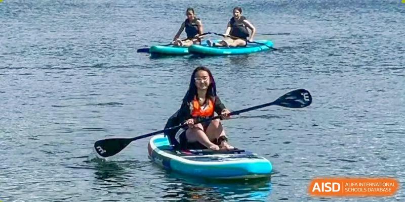 A student sits on a paddle board in the water, holding a paddle, with other students in the background. Suzhou Science and Technology Town Foreign Language High School.