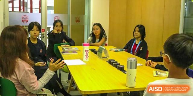 Older students at Dehong Beijing International Chinese School are engaged in a discussion or small group meeting around a yellow table.