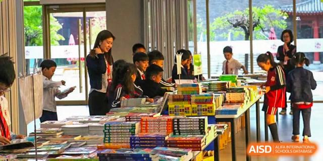 Students at Dehong Beijing International Chinese School browsing and reading books at a book fair or library area with a teacher.