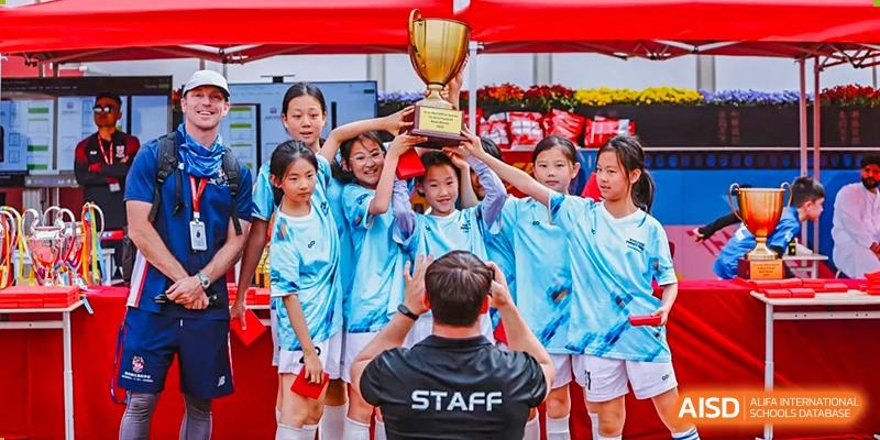 Students at Dehong Beijing International Chinese School celebrating a victory, holding a large trophy while posing with a staff member.