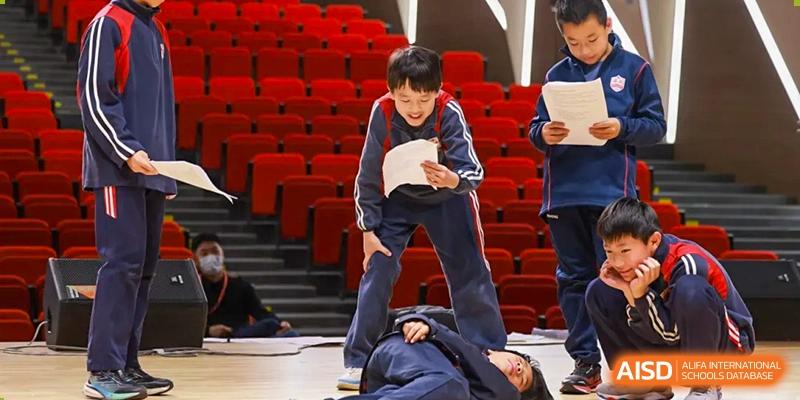 Students at Dehong Beijing International Chinese School performing a dramatic or acrobatic routine on a stage with red seating.