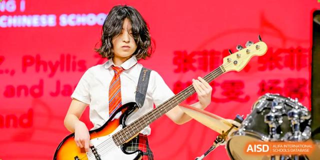 A student at Dehong Beijing International Chinese School passionately playing an electric bass guitar on a brightly lit stage.