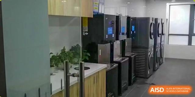 A neat row of black, modern dispensing machines is installed on a counter within a brightly lit, glass-enclosed interior kitchen or service area, at Cambridge International School Litai College. SISU.