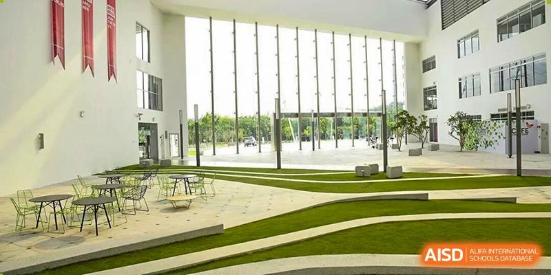 A modern building interior features a tiered, wavy landscape of green artificial turf extending towards a patio seen through tall glass windows, at Cambridge International School Litai College. SISU.