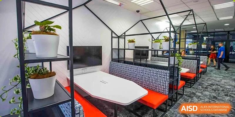 An indoor, enclosed seating booth features a distinctive black architectural frame, a long white table, and bright orange benches with a decorative blue-tiled base, at Cambridge International School Litai College. SISU.